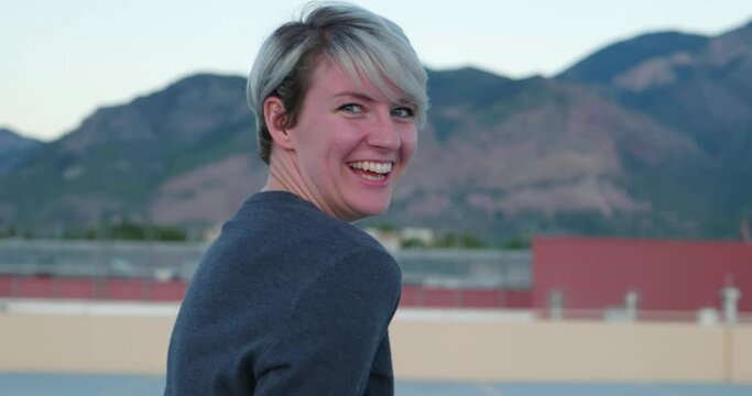 Happy silly and playful caucasian white girl looking into camera and laughing smiling, with mountains and buildings in background. In Cinema 4K (4096x2160) 30fps slowed from 60fps.
