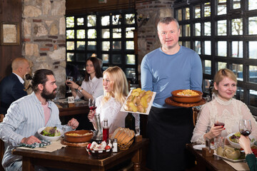 Hospitable owner of cosy country restaurant meeting guests with traditional meals in hands