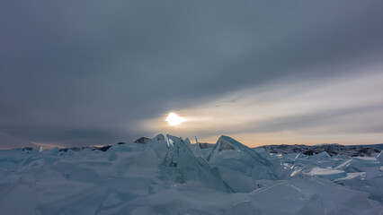 On a frozen lake - a block of turquoise ice hummocks. The glare of the setting sun on the edges. Orange glow in the evening cloudy sky. Baikal
