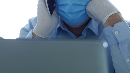 Doctor in a Hospital Office with a Face Mask and Protective Gloves Use Smartphone in Front of the Laptop Screen