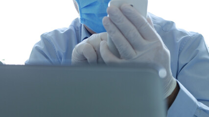 Doctor in a Hospital Office with a Face Mask and Protective Gloves Use Smartphone in Front of the Laptop Screen