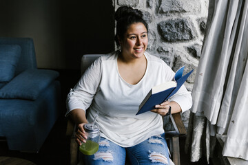 Portrait of curvy latin girl holding a jar of green juice while reading a book at home