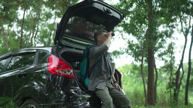Asian Senior man, retired, sitting on a car use binoculars looking at the birds in the trees, in the woods, and writing down the information in a notebook. happily on vacation days
