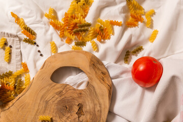 One tomato, a fragment of a cutting board, multi-colored pasta. 