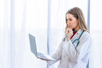 female doctor with stethoscope working with laptop computer and writing on paperwork on wooden table in Hospital background