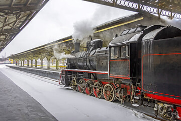 Steam locomotive stands on the platform of the station, winter cold snowy day.