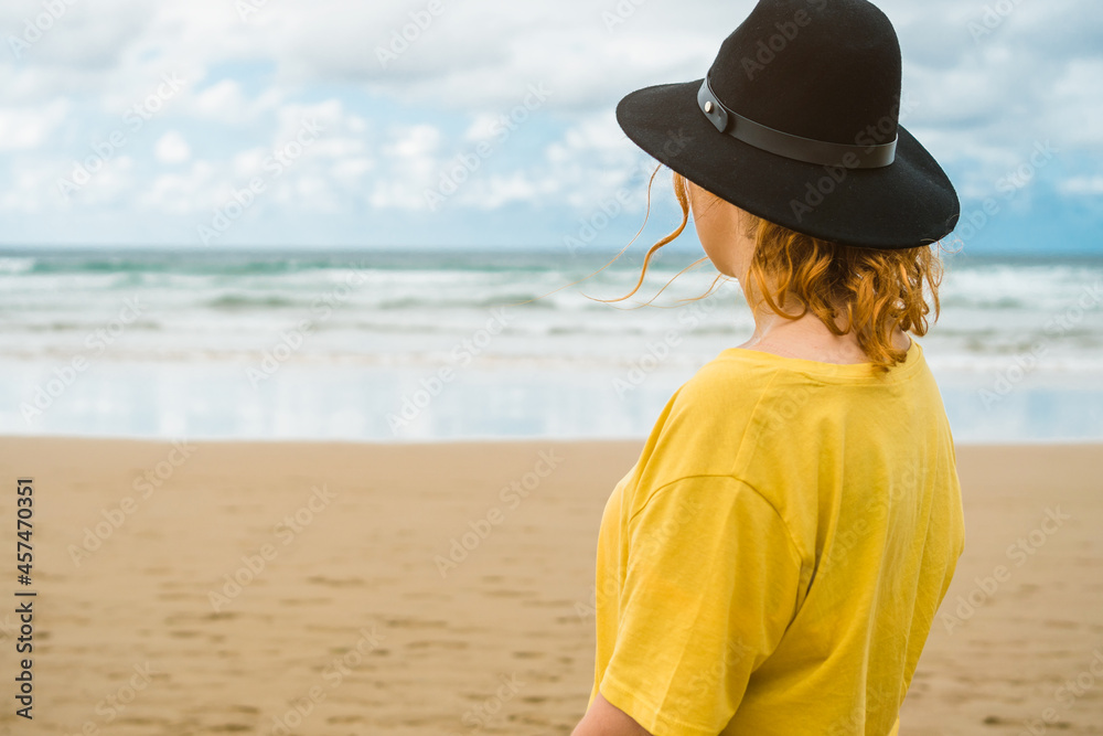 Wall mural closeup of unrecognizable woman in yellow shirt and black hat standing on the beach looking at the s