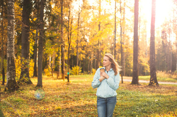 A beautiful happy blonde woman walks through the autumn park with a cup of warming drink in her hands. Walk on a sunny autumn evening. Healthy lifestyle.