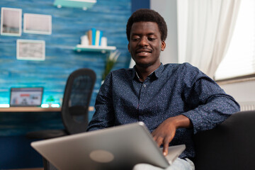 Black student sitting on couch in living room studying management graph for univeristy course using school platform on laptop computer. Teenager working at financial project. Computer user