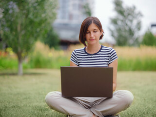 happy asian female student working with laptop in modern university campus park. woman in striped T-shirt and light trousers works at gray laptop. The student studies outdoors.