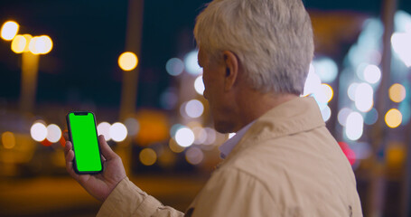 Bokeh shot of aged man holding smartphone with blank green screen outdoor
