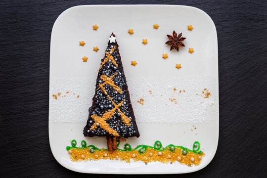 Weihnachtliches Bild auf einem weißem Teller: Brownie als Weihnachtsbaum dekoriert mit schwarzem Zuckerguss, Sternen, Zuckerperlen, Puderzucker und goldenem Zuckerstaub