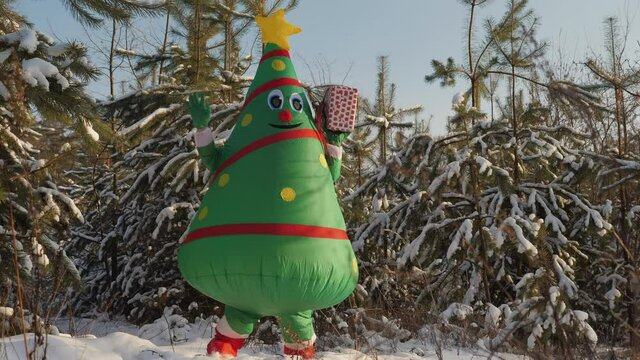 A man in an inflatable costume with a Christmas tree doll dances cheerfully in a pine forest with a gift box in his hand