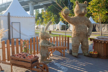 Fairy-tale characters bear and hare made of straw, against the background of a cart with carrots...
