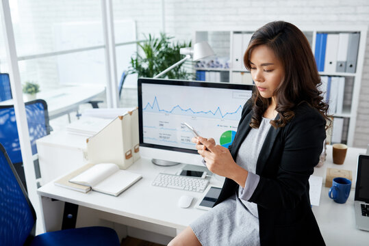 Serious businesswoman sitting on office desk and reading important text messages from colleagues who work from home