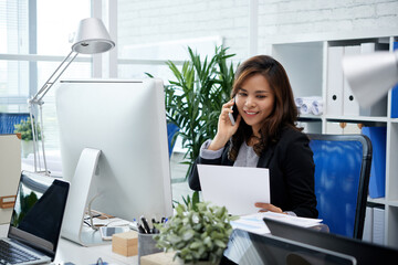 Pretty smiling female entrepreneur talking on phone with colleague from another department when...