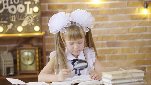 Little girl reads a book with magnifier, sitting at table on background of a school class. An elementary school girl with white bows. Holiday of the first of September, the day of knowledge.