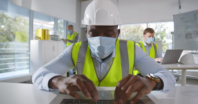 African engineer wearing protective mask working on laptop in construction office