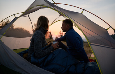 Girl sitting in tent, covered with sleeping bag, with her guy, holding metal cup and enjoying the...