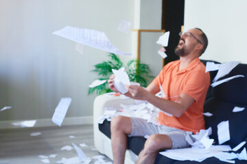 A young hipster man with a beard sitting on the sofa at home throwing papers in the air while laughing out loud due to stress