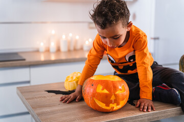 Child sitting at kitchen table dressed as pumpkin playing with decorated halloween pumpkin. Happy Halloween