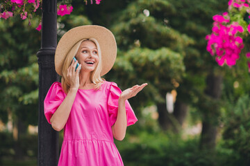 Photo of adorable impressed lady pensioner dressed pink clothes headwear walking talking modern device smiling outdoors outside city street