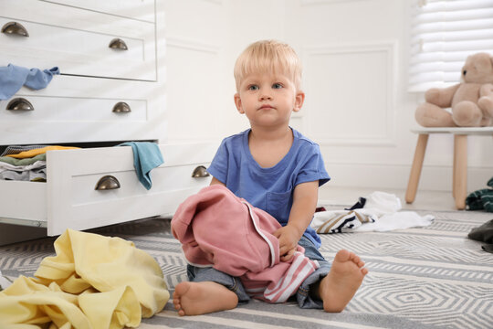 Cute Little Boy Playing With Clothes Near Dresser On Floor At Home