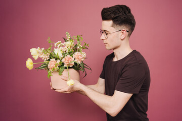 A young man is holding a beautiful bouquet of flowers in his hands on a pink background. The guy decided to make a surprise to his girlfriend. Mother's Day. Women's day. Flowers delivery.