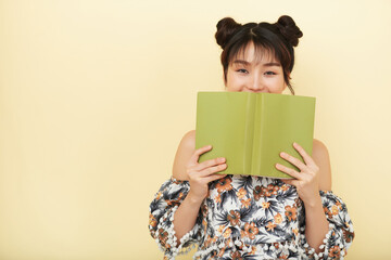 Happy pretty young Vietnamese woman hiding her smile behind opened book and looking at camera