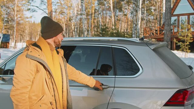 Handheld tracking of cheerful bearded man, his wife and excited little daughter getting out of car parked in snow before cozy A-frame cabin in forest, then unloading trunk