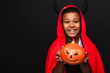 cheerful african american boy in devil halloween costume holding carved pumpkin isolated on black
