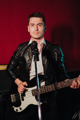 Talented handsome young guitarist man singing a song in studio recording on red background surrounded by instruments. Passion, hobby, singer, electric guitar