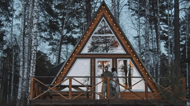 Medium shot of happy couple walking out of cozy A-frame cabin in woods and enjoying scenery while standing wrapped in warm blanket on their front porch