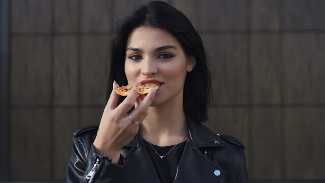 Portrait happy young brunette woman in trendy stylish look wearing black leather jacket eat slice of pizza looking at camera and smiling standing by wall in city street. Eating snack outdoors