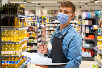 Young store worker man in a medical mask wearing apron black color holding the opened journal and a pen in his hands, doing the inventory in the store.