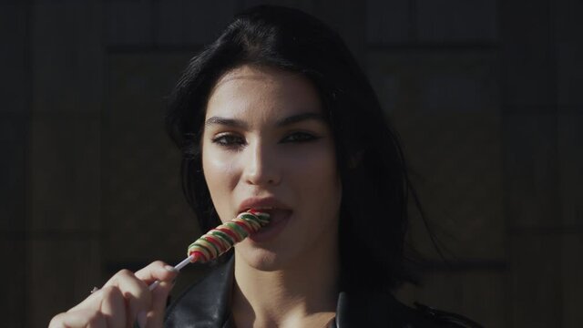 Portrait happy young brunette woman in trendy stylish look wearing black leather jacket seductively licks colorful lollipop candy looking at camera and smiling standing by wall in city street