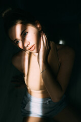 Naklejka premium A beautiful woman is standing at the window at home and enjoying the sunrise. The woman smiles, closing her eyes and rejoicing in the new day. Woman wearing a men's blue button-down