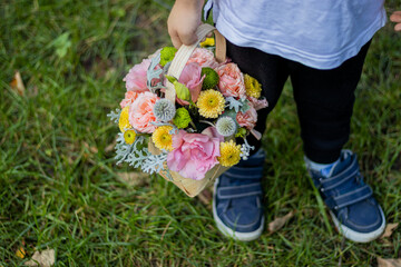 A little kid is holding a basket of beautiful, mixed flowers in his hands at the bottom of his legs. the boy is in the park, summertime.