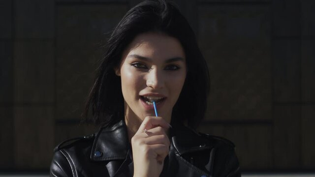 Portrait happy young brunette woman in trendy stylish look wearing black leather jacket seductively licks colorful lollipop candy looking at camera and smiling standing by wall in city street