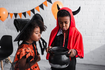 african american boy in halloween costume holding witch cauldron with potion near sister in pointed...