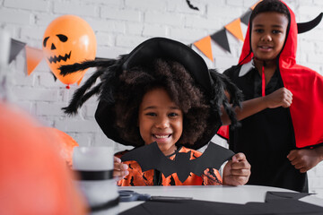 happy african american girl in halloween costume holding paper cut bat near brother - Powered by Adobe