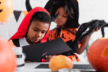 african american boy cutting black paper near sister in witch halloween costume