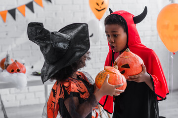 spooky african american kids in halloween costumes holding carved pumpkins at home