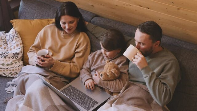 High angle shot of happy mother, father and little girl sitting under blanket and watching movie on laptop in their cabin on cold winter or autumn day