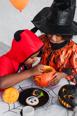 african american kids in halloween costumes carving pumpkin near cookies