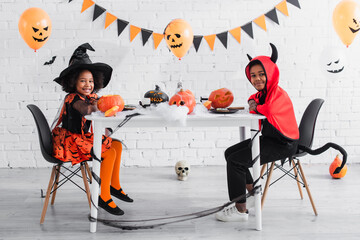 happy african american kids in halloween costumes sitting at table with pumpkins and decoration