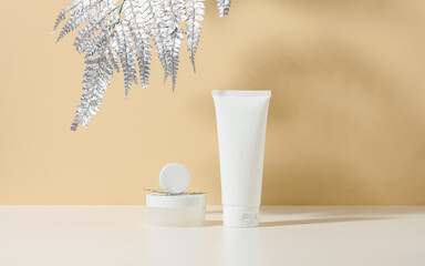 white tube for cosmetics, a jar of cream and silver leaf on a white table. Cosmetic on a beige shadow backgroun