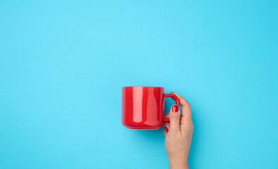 red ceramic cup in a female hand on a blue background, drink and hand are raised up, coffee break