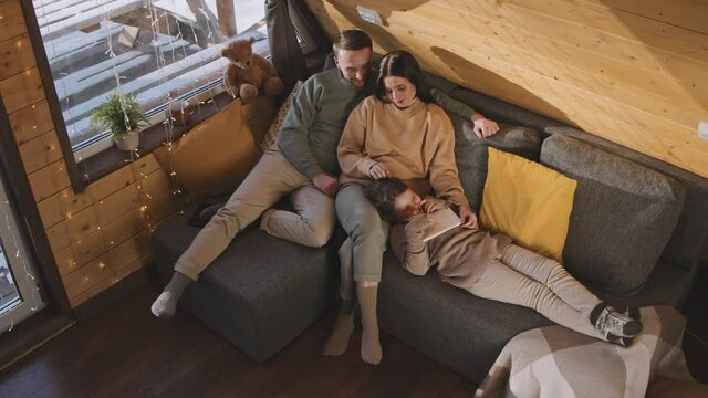 High angle shot of cheerful parents and cute little girl relaxing together on couch in cozy cabin in winter. Girl lying on mothers lap and holding tablet