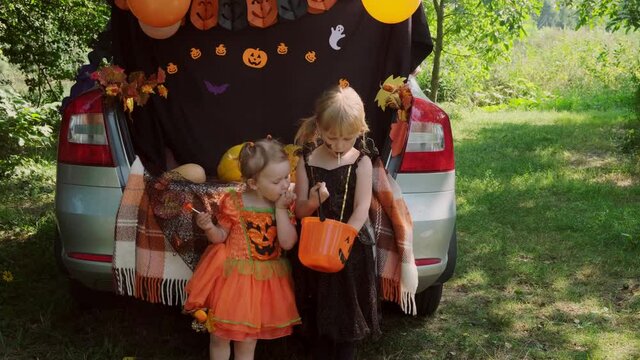 Two little sisters celebrating Halloween and eating candy from buckets sitting in trunk of car.